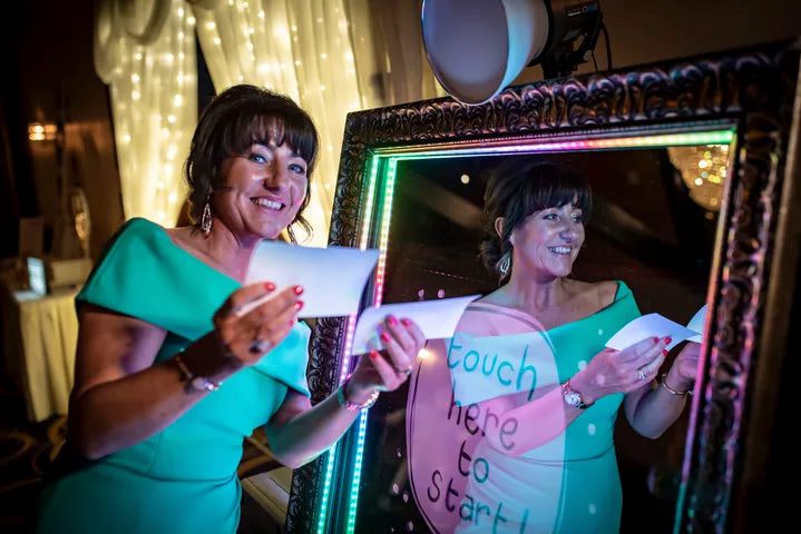 Smiling Woman in a Stylish Green Dress Posing in Front of the Magic Mirror Photo Booth, Holding Memorable Photos